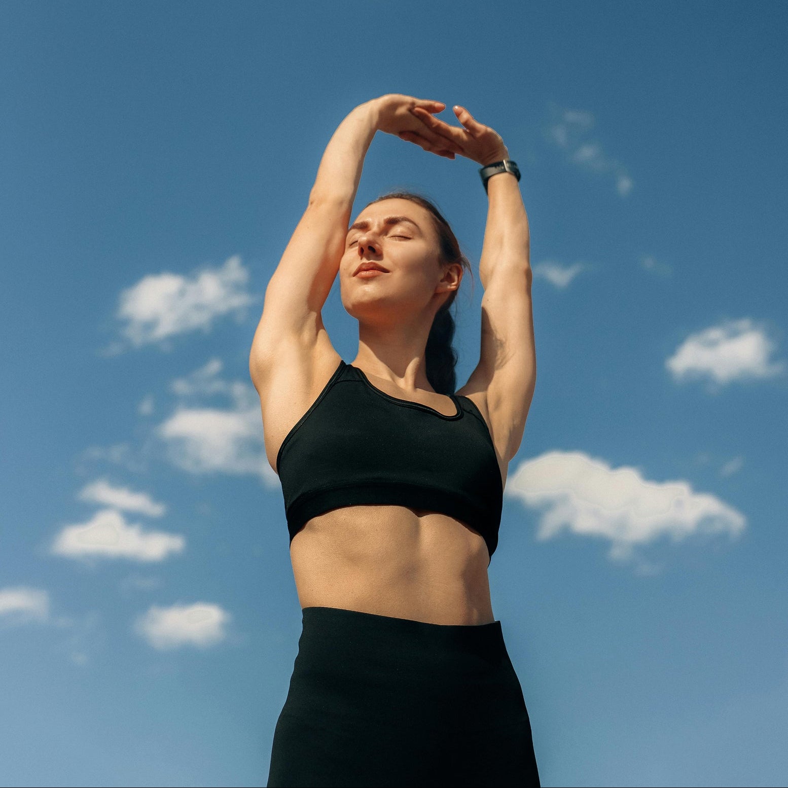 Woman in black athletic wear stretching against a blue sky with scattered clouds
